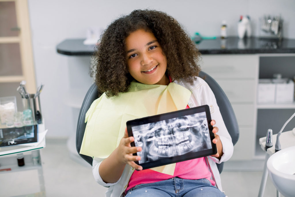 A smiling mixed-race girl with curly hair sits in a dental chair, holding a digital tablet displaying dental x-rays of her teeth in a pediatric dentistry setting.